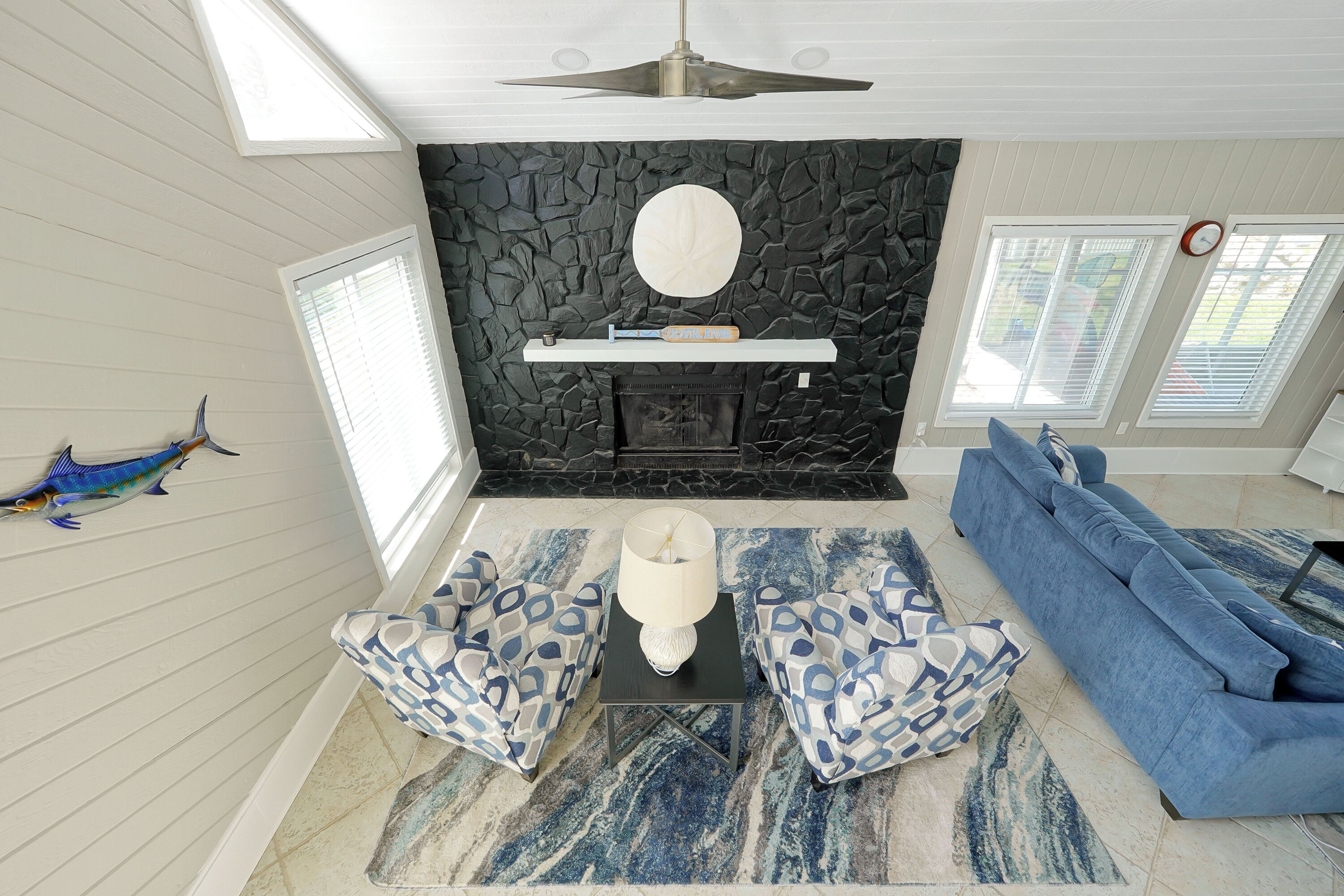A living room with a stone fireplace, blue patterned sofas, and a ceiling fan.