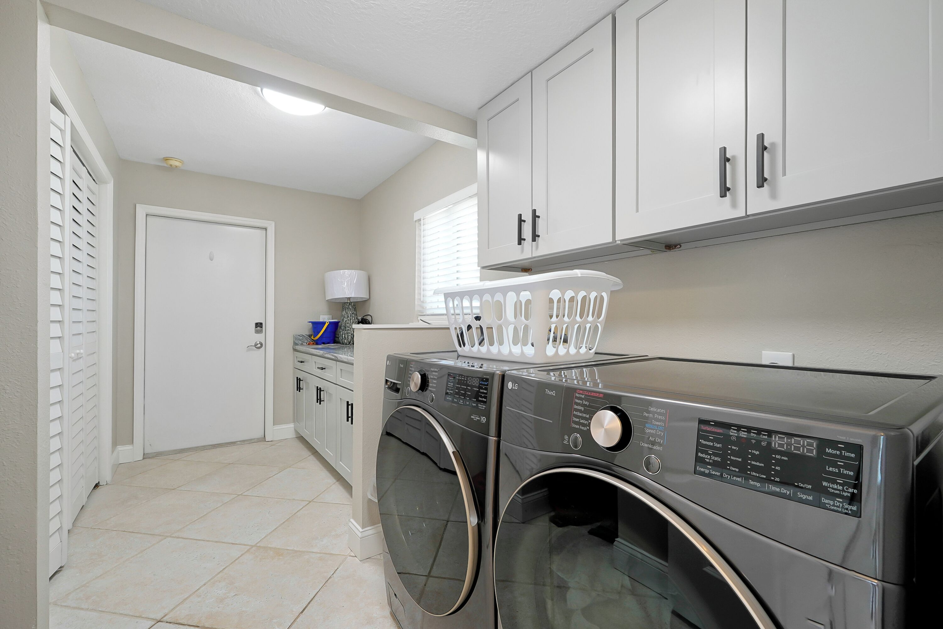 A laundry room with two front-loading washing machines, a white door, and a window with blinds.