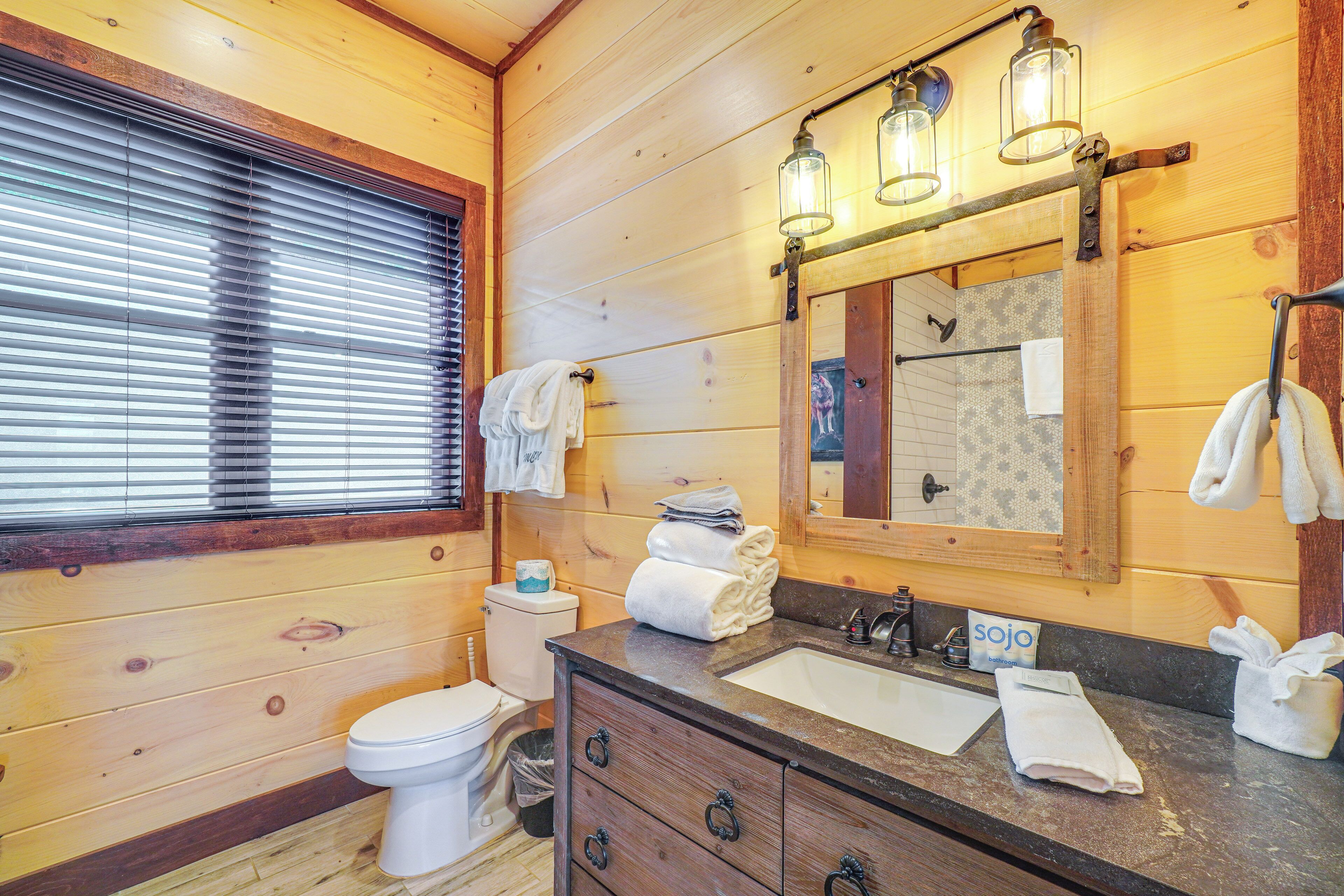 A wooden cabin bathroom with a toilet, sink, and mirror. There are towels hanging on the wall and a window with blinds.