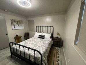 Une chambre à coucher comprenant un lit, une table de chevet, une chaise, une petite table et une œuvre d’art accrochée au mur.