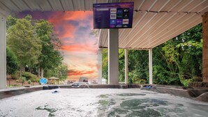 A hot tub area with a digital screen displaying various streaming services, surrounded by lush greenery and a sunset view. - Heated Pool, Hot Tub, Game Rm, Boat Dock, Lakefront Views (Lake Norman of Catawba)