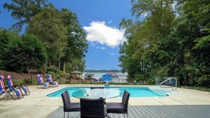 A swimming pool with lounge chairs and a table on a wooden deck, surrounded by trees and a view of a lake. - Heated Pool, Hot Tub, Game Rm, Boat Dock, Lakefront Views (Lake Norman of Catawba)