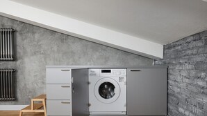 Un lavadero pequeño con lavadora y secadora integradas, un taburete de madera y un radiador.
