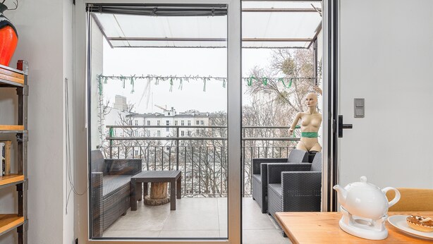 Una sala de estar moderna con un ventanal corredera que da a un balcón. El balcón tiene muebles de mimbre, una mesita pequeña y una tetera blanca sobre una mesa de madera.