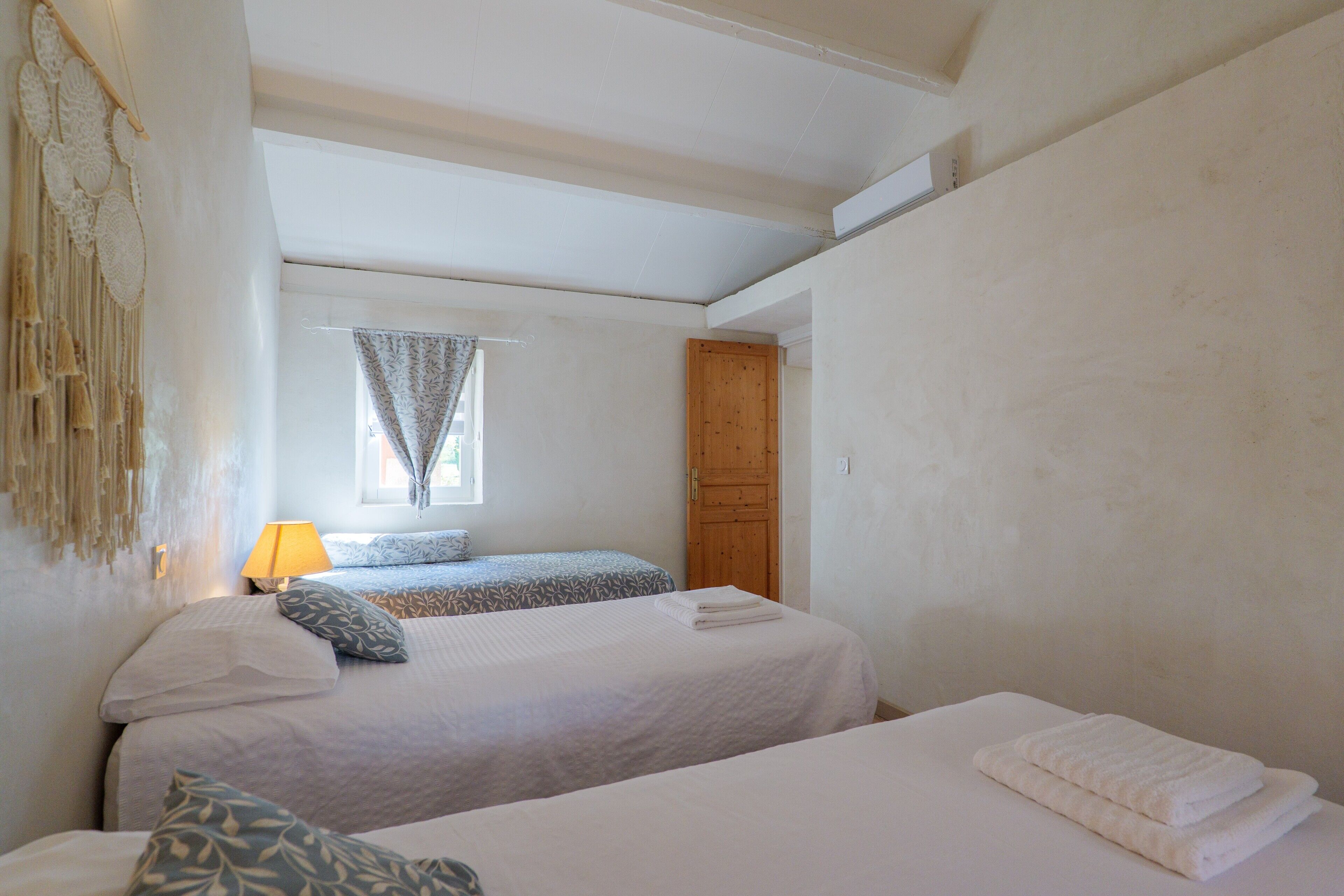 A bedroom with two beds, a window with curtains, a wooden door, and a wall-mounted macrame decoration.