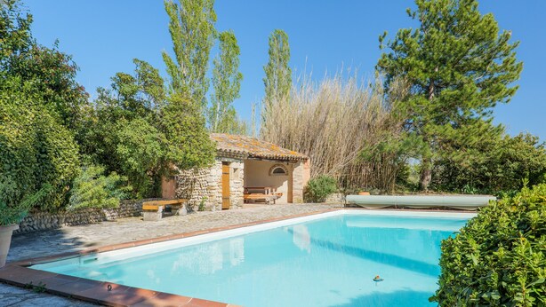 Une piscine aux bords recouverts de pierres, entourée de verdure et bordée d’un bâtiment en pierre sur lequel se trouve un banc.