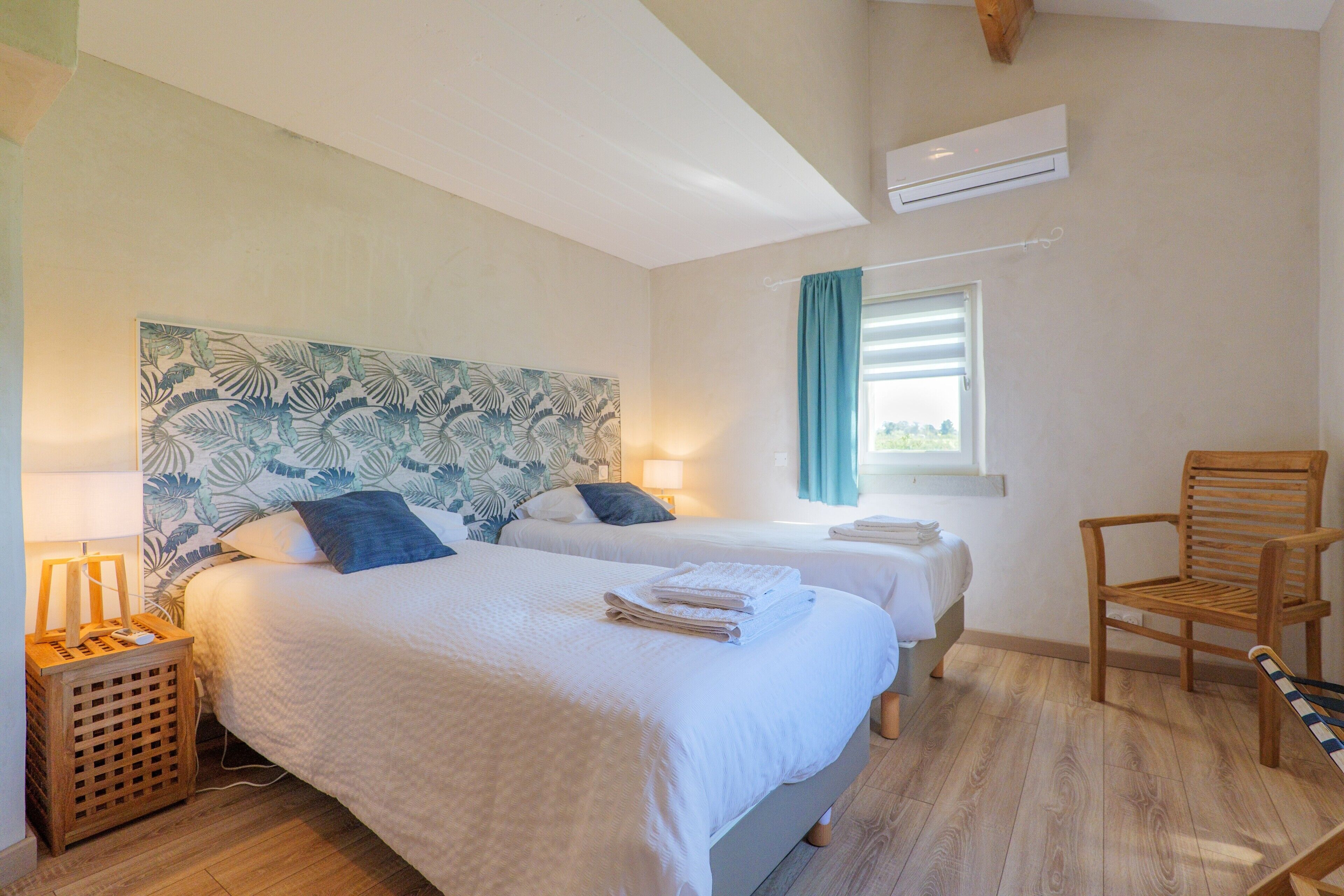 Une chambre d’hôtel avec deux lits, une chaise en bois, une fenêtre avec des rideaux et un appareil de climatisation.