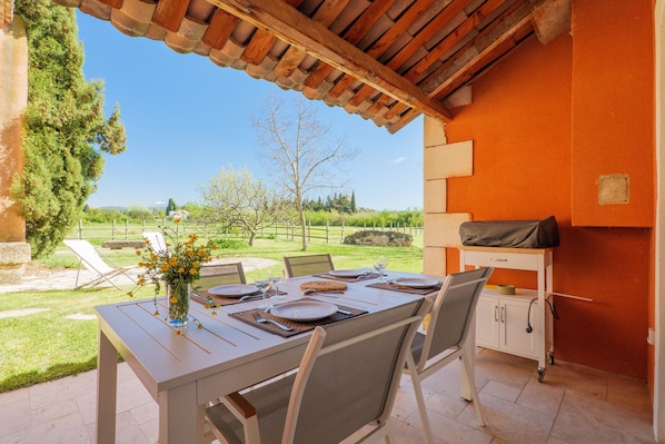 An outdoor dining area with a table set for four, chairs, a vase with flowers, and a grill on the right.