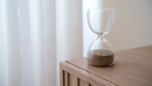 Eine Sanduhr auf einem Holztisch, im Hintergrund ein weißer Vorhang.
