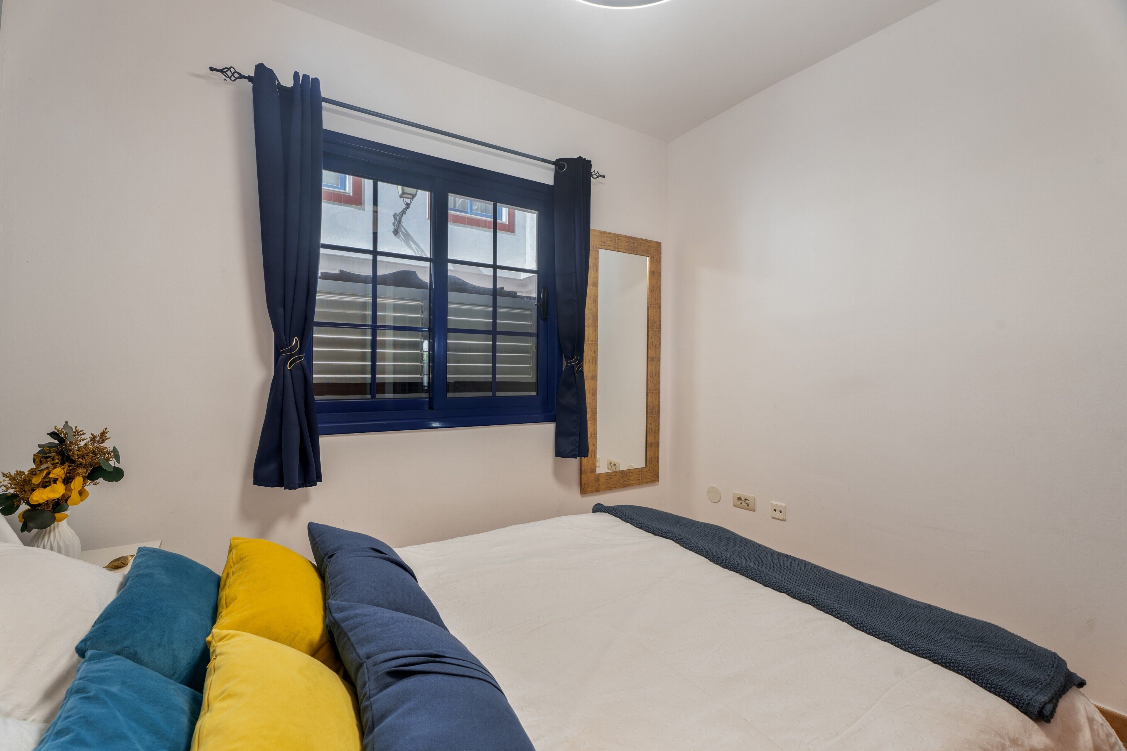 Een slaapkamer met een bed, een raam met gordijnen, een spiegel en een vaas met bloemen.