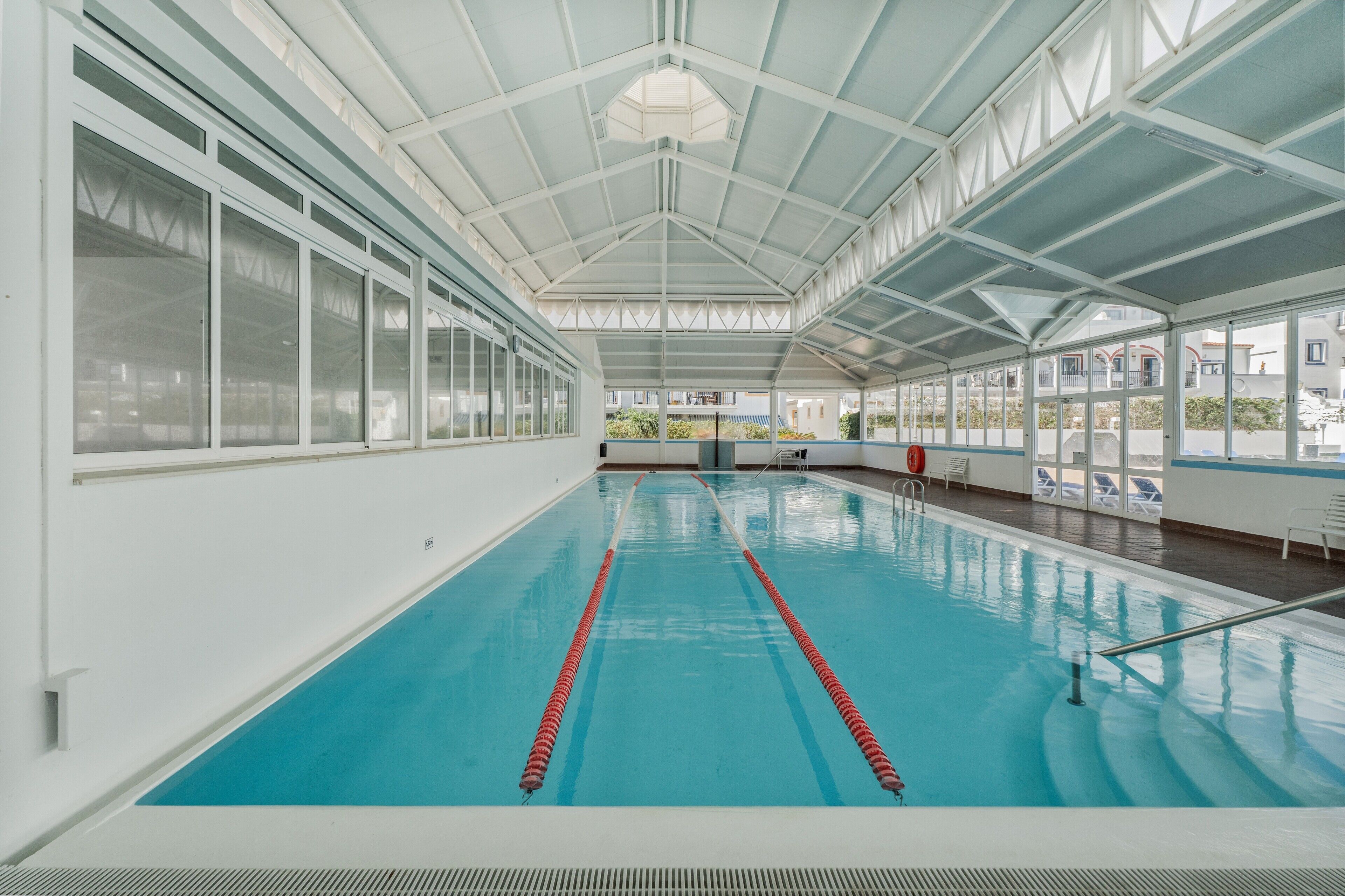 Piscina interna com paredes de vidro transparente, teto alto e vista para edifícios externos.