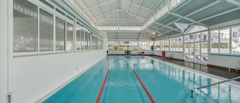 Piscina interna com paredes de vidro transparente, teto alto e vista para edifícios externos.