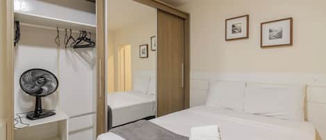 A hotel room with a bed, a fan, a wardrobe, and two framed pictures on the wall.