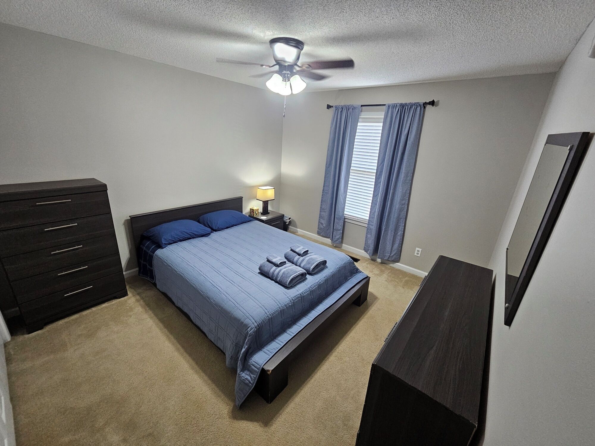 Une chambre à coucher comprenant un lit, une commode, une table de chevet, une lampe et un ventilateur de plafond.