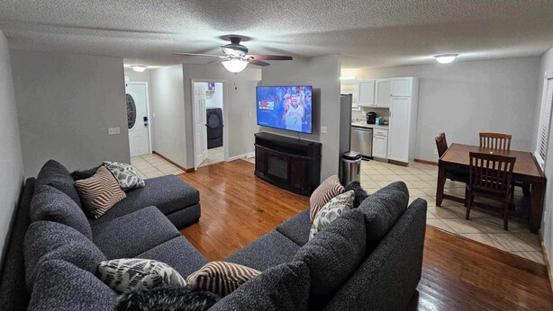 A living room with a gray sectional sofa, a TV, and a ceiling fan. There is a dining area with a wooden table and chairs.