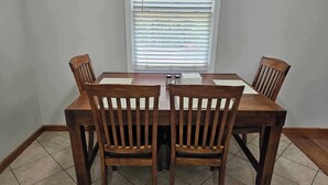 A dining table with four wooden chairs, a window with blinds, and a tiled floor.