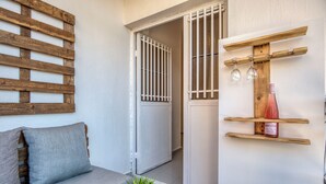 A room with a wooden bench, a grey sofa, a small table with a plant, and a wall-mounted shelf with a bottle and glasses. - Agrius - Elegant Studio & Balcony in Batroun (Batroun)