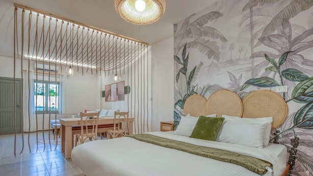 A bedroom with a bed, a desk, and a chair. There is a window with curtains and a wall with a tropical plant mural. - Agrius - Elegant Studio & Balcony in Batroun (Batroun)