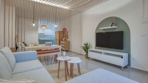 A modern living room with a sofa, a TV on a white stand, a dining area with a table and chairs, and a kitchen in the background. - Agrius - Elegant Studio & Balcony in Batroun (Batroun)
