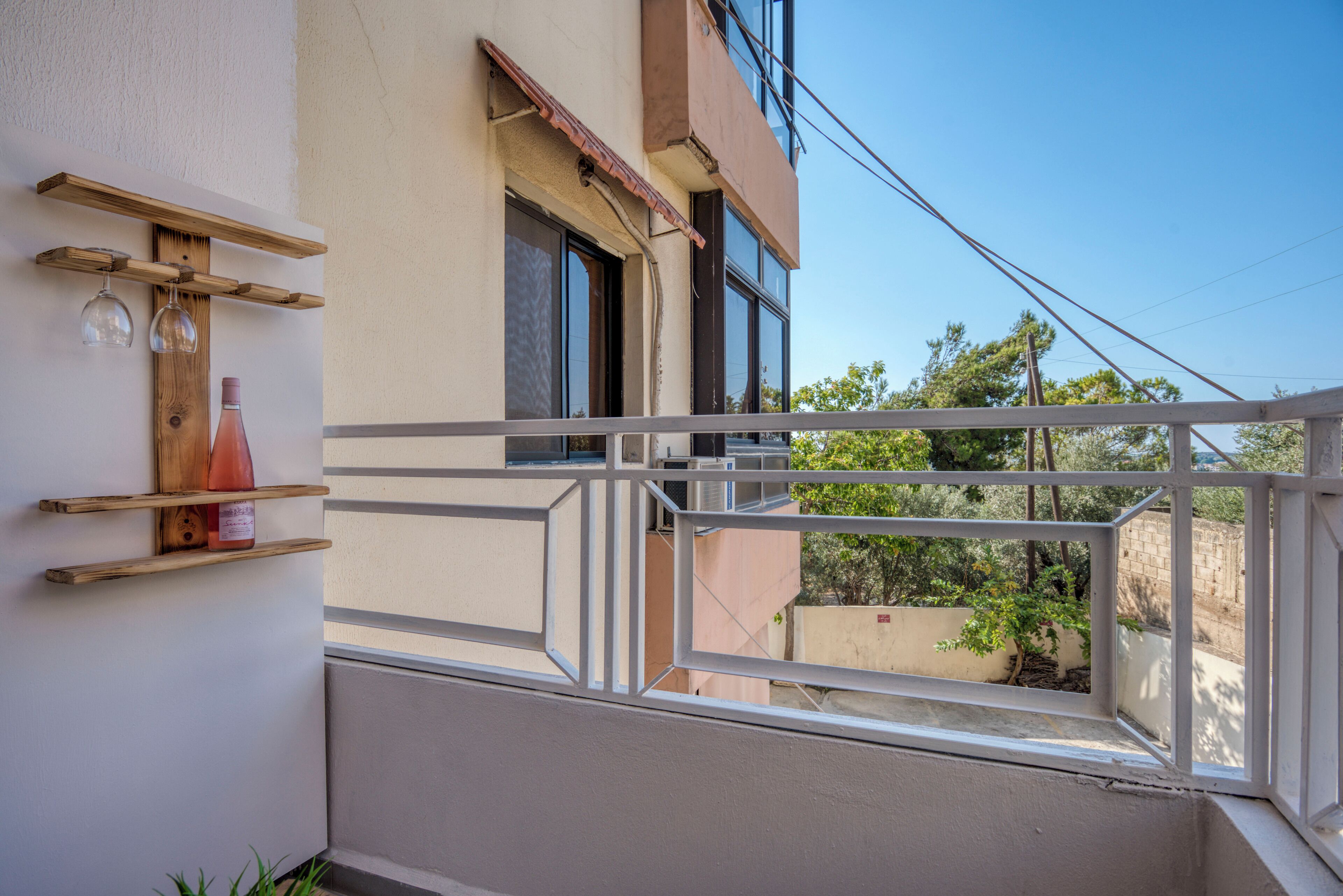 A balcony with a wooden shelf holding a bottle and glasses, a view of a building, and a clear sky.