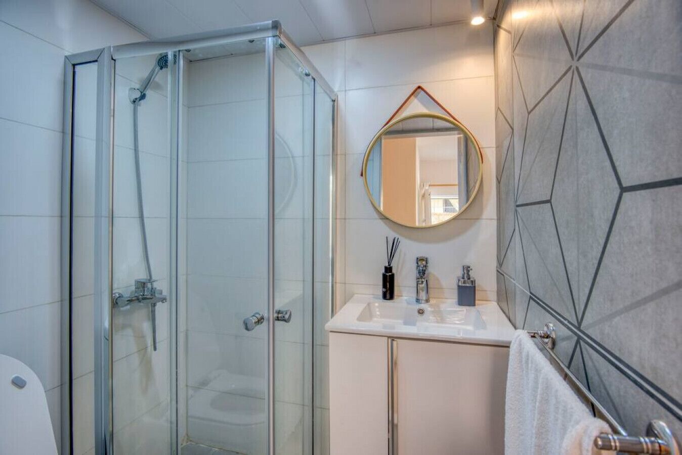 Un baño moderno con cabina de ducha de vidrio, un espejo redondo y un lavamanos blanco con un salpicadero de azulejos geométricos.