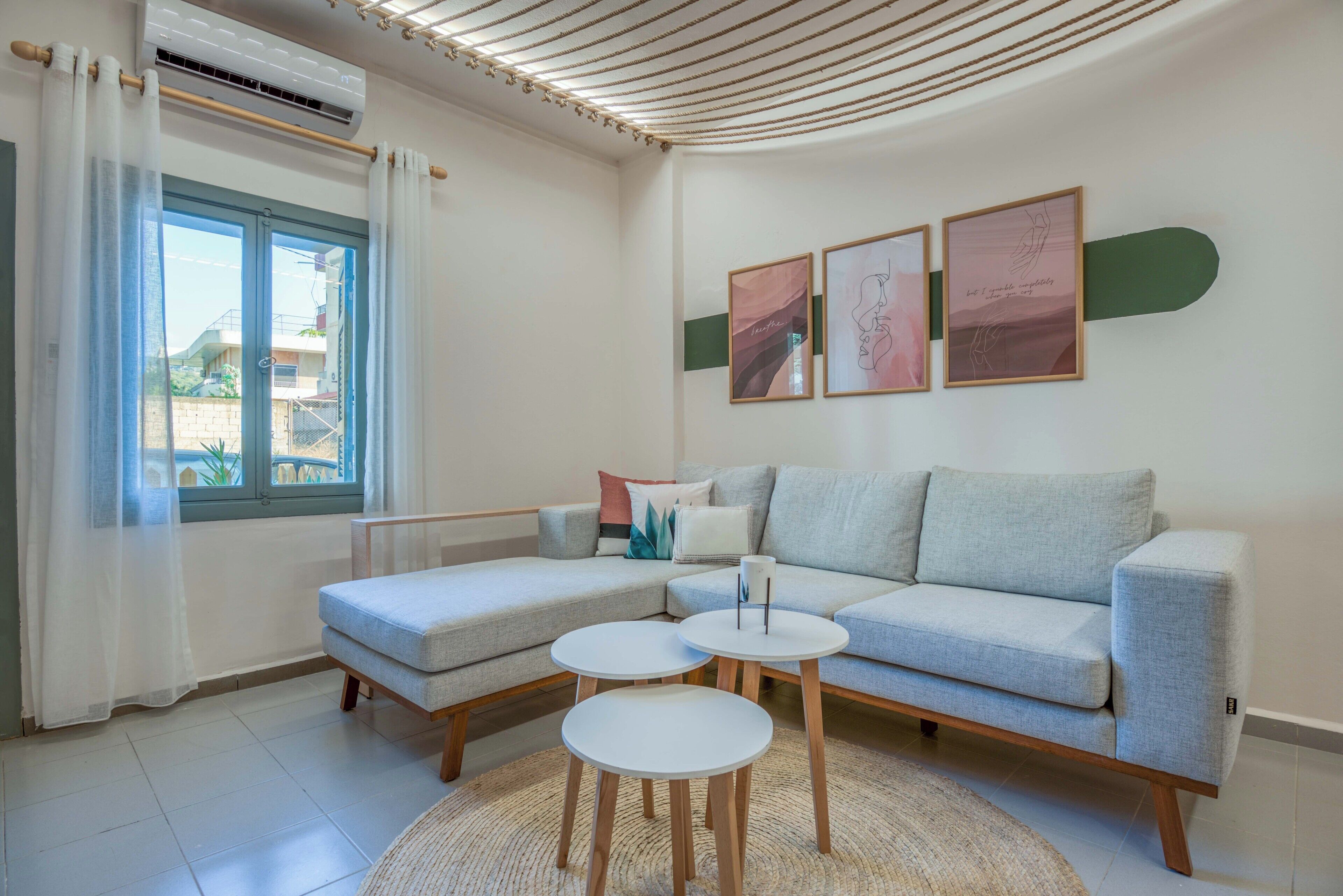 Een moderne woonkamer met een grijze modulaire bank, twee ronde witte tafels, een groot raam met dunne gordijnen en drie ingelijste kunstwerken aan de muur.