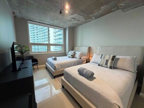 A bedroom with two beds, a desk with a computer, and a large window with a view of high-rise buildings. - Midtown Miami Condo - Walk Wynwood - We Host (Miami)