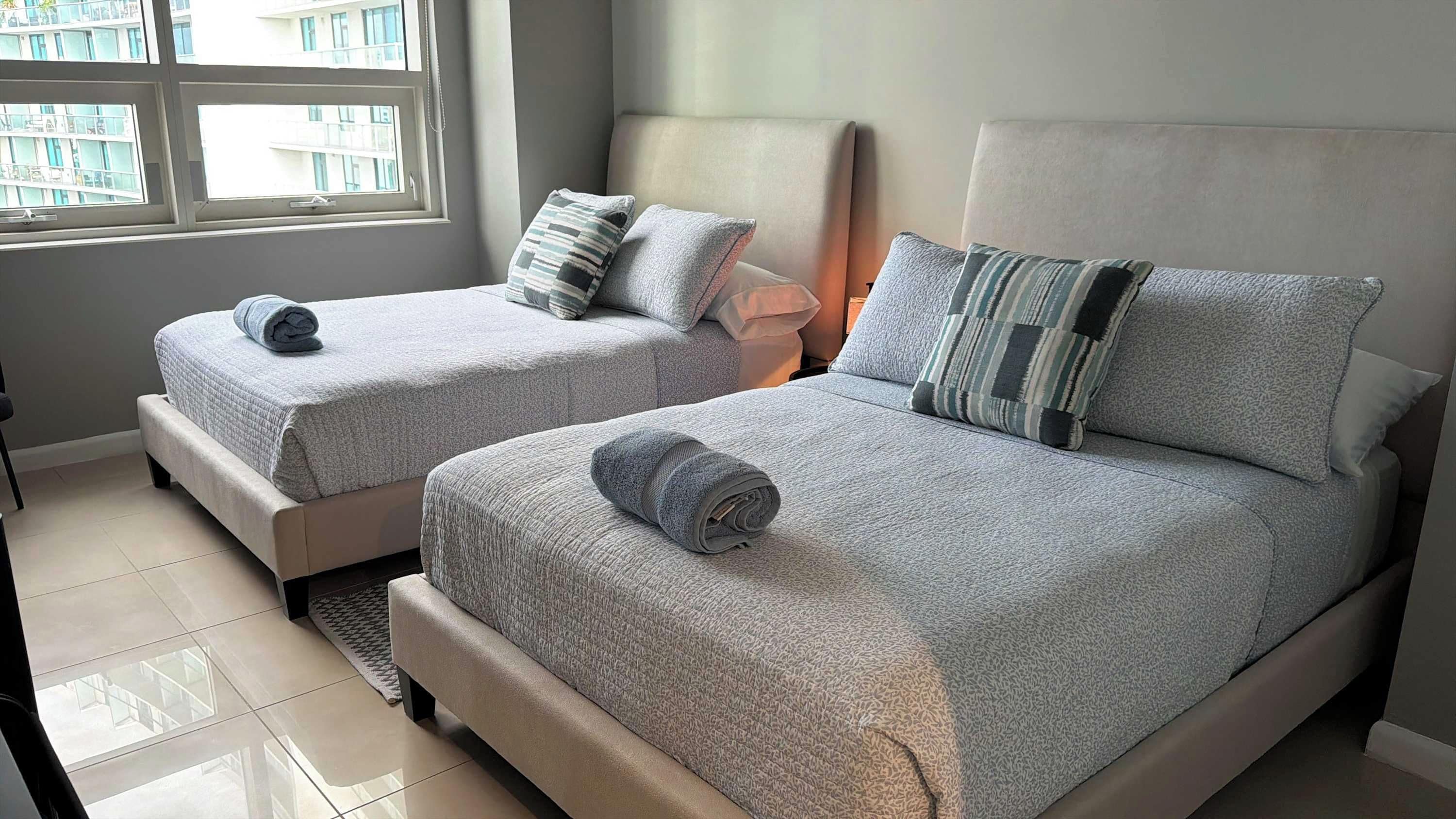 Dos camas con ropa de cama gris, una ventana con vistas a edificios y un suelo de baldosas.