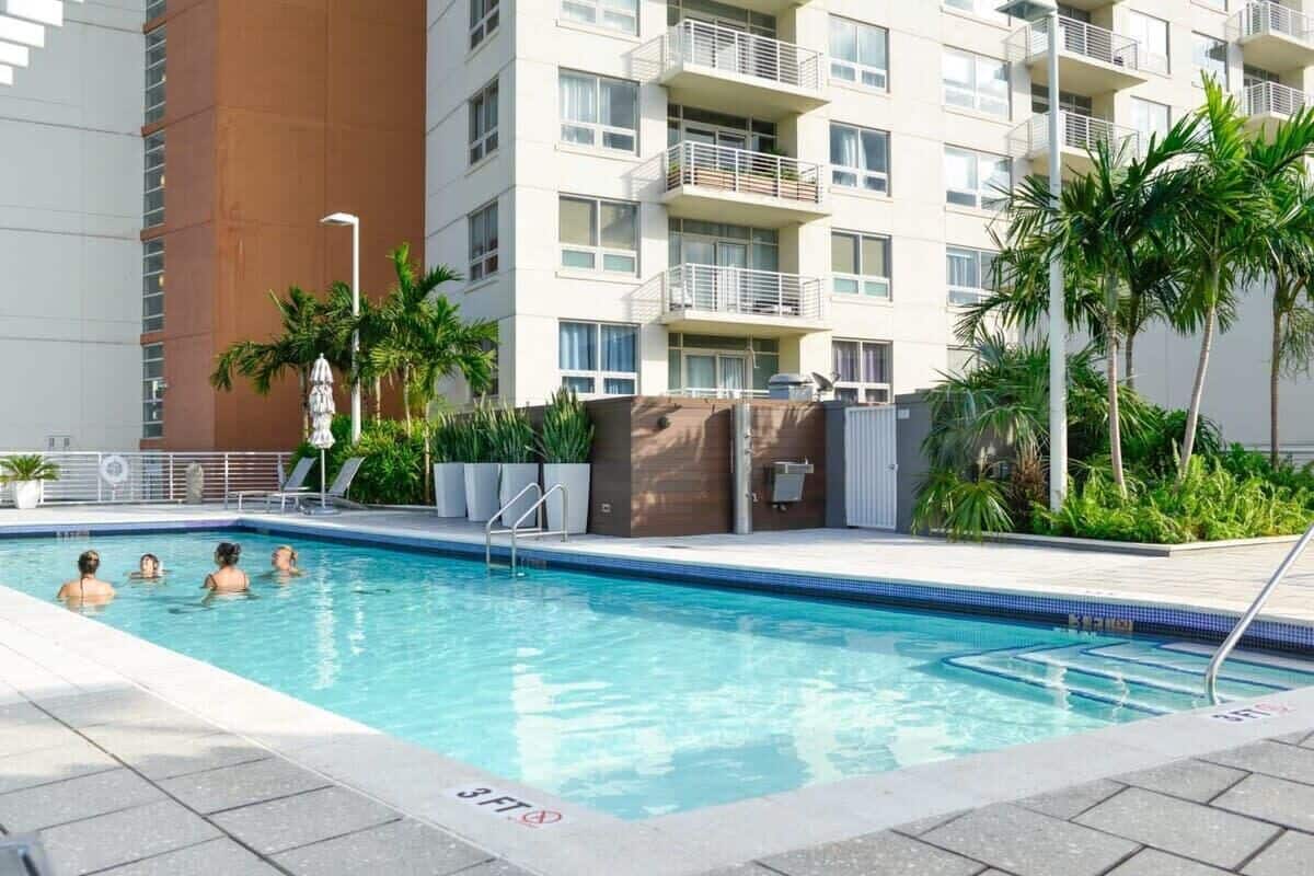 A swimming pool with people, surrounded by a modern building with balconies and palm trees.