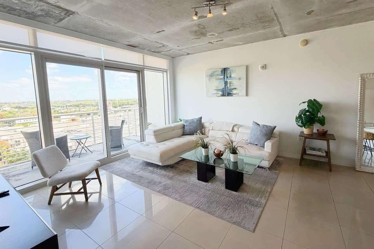 A modern living room with a sofa, armchairs, a glass coffee table, and a large window offering a city view.