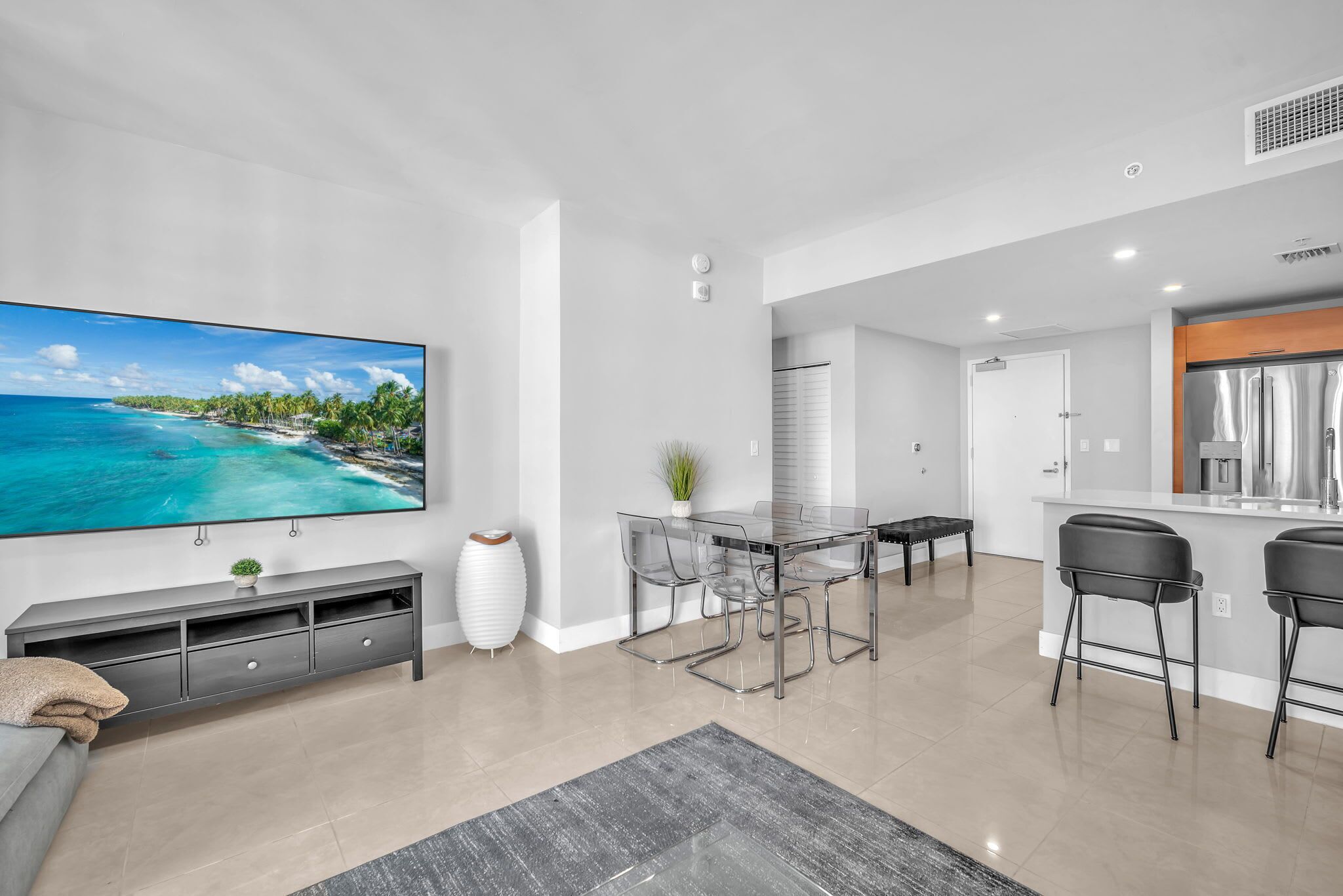 A modern living room with a large flat-screen TV, a glass dining table with chairs, a sofa, and a kitchen area with a refrigerator and bar stools.