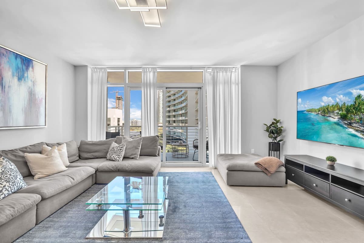 A modern living room with a glass coffee table, grey sofas, a large TV, and a view of a high-rise building.