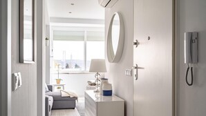 Ein modernes Hotelzimmer mit grauem Sofa, einem Nachttisch mit Lampe, einem Spiegel und Blick auf die Stadt durch große Fenster.