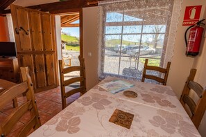 Ein Essbereich mit einem Tisch für zwei Personen, Holzstühlen, einem Fenster mit Blick auf parkende Autos und einen Hügel, sowie einem an der Wand befestigten Feuerlöscher.