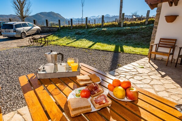 Une table en bois dressée pour le petit-déjeuner, avec du pain, du jambon, des tomates et des fruits, et en arrière-plan, une vue imprenable sur les montagnes.