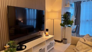 Un salon moderne doté d’un grand téléviseur à écran plat, d’un meuble de rangement blanc, d’une lampe sur pied, d’une plante en pot et d’un canapé agrémenté d’un plaid.
