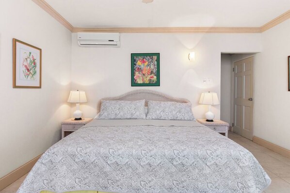 A bedroom with a large bed, two bedside tables with lamps, a wall-mounted air conditioner, and framed botanical artwork.