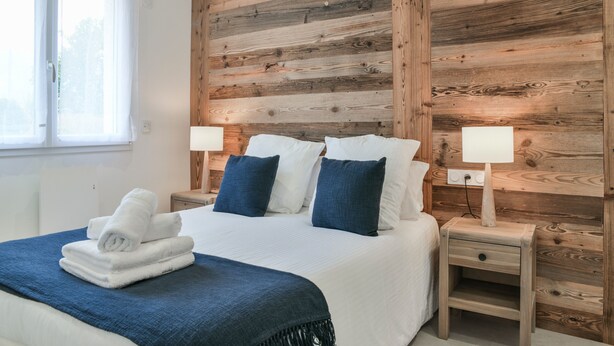 Una camera da letto con parete in legno, un letto con biancheria da letto bianco e blu, due comodini con lampade e una finestra con tende.