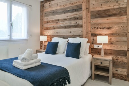 A bedroom with a wooden wall, a bed with white and blue bedding, two bedside tables with lamps, and a window with curtains.