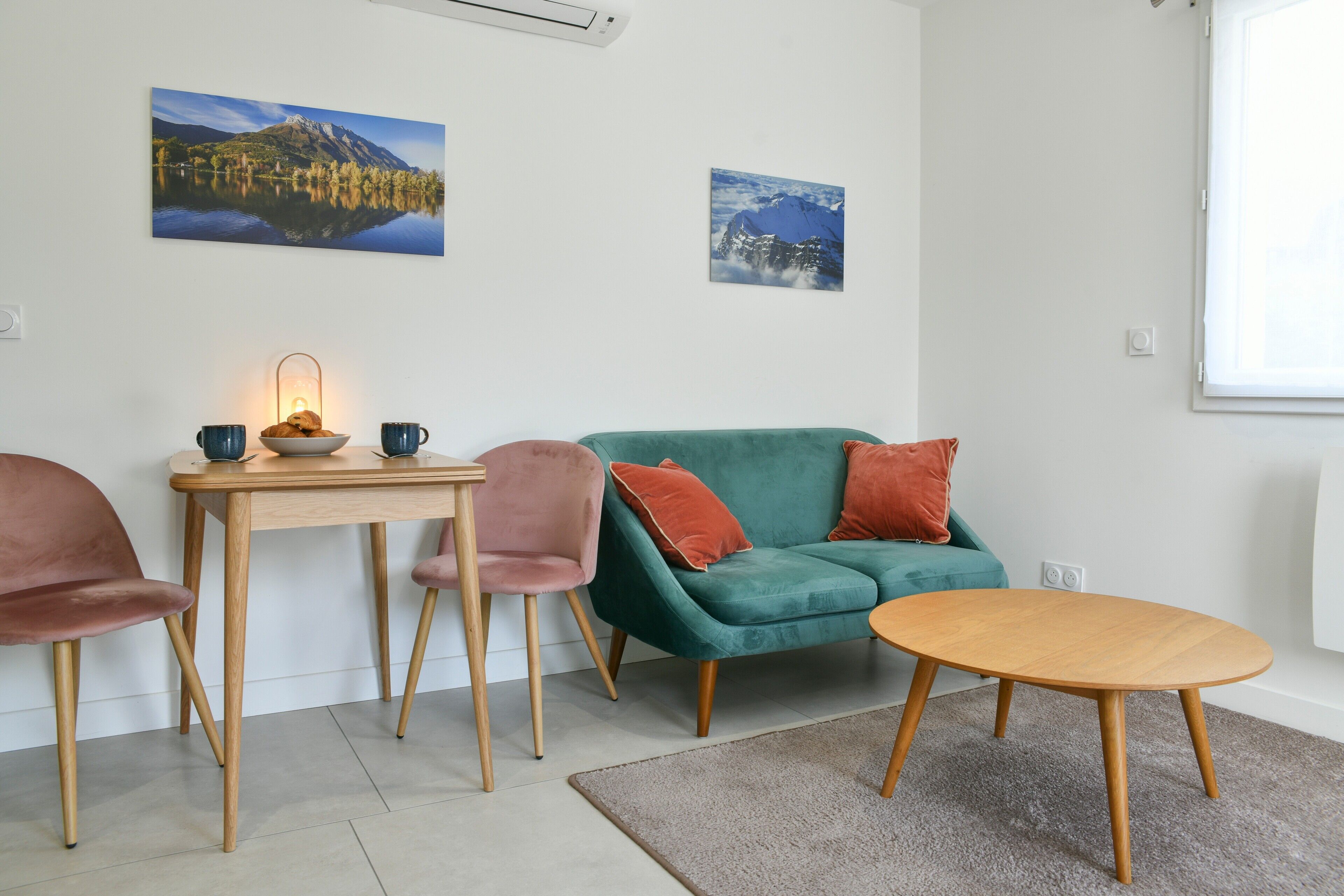 A modern living room with a green sofa, a wooden table, and two chairs. There are two wall-mounted pictures depicting mountains and a lake.