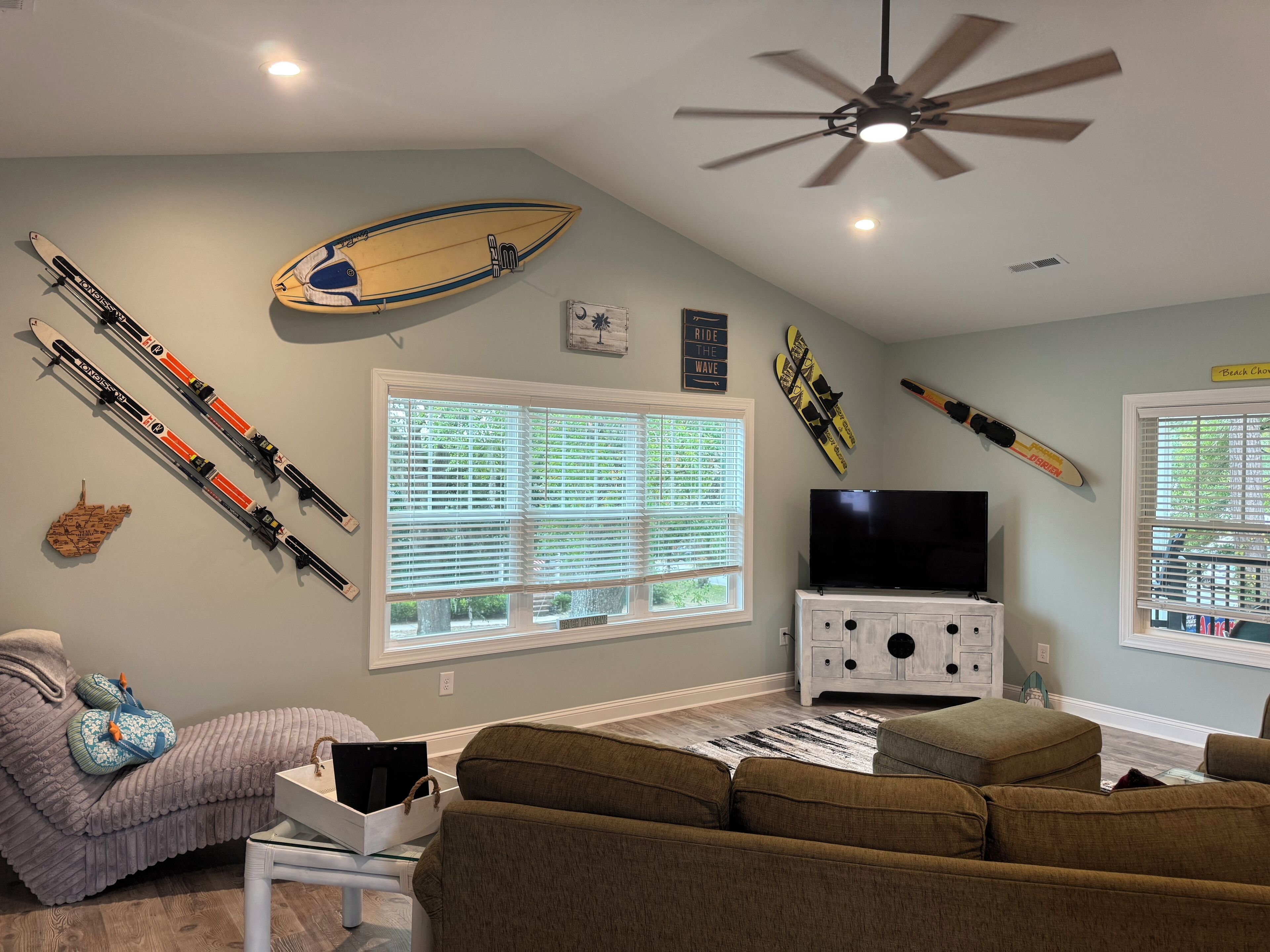 Una sala de estar con ventilador de techo, televisión y esquíes y tablas de snowboard en la pared.