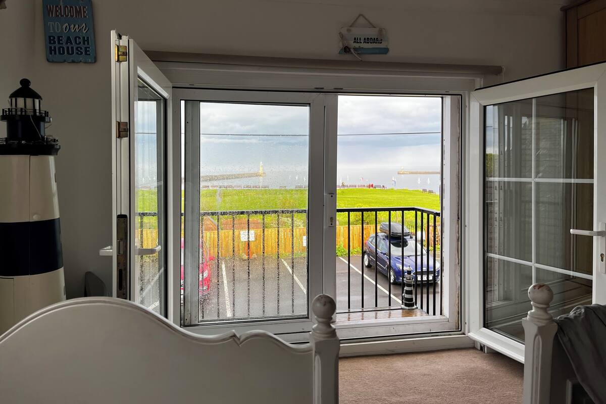 A room with a large sliding glass door leading to a balcony with a view of a grassy area and a body of water. There is a sign on the wall that reads "WELCOME TO OUR BEACH HOUSE."