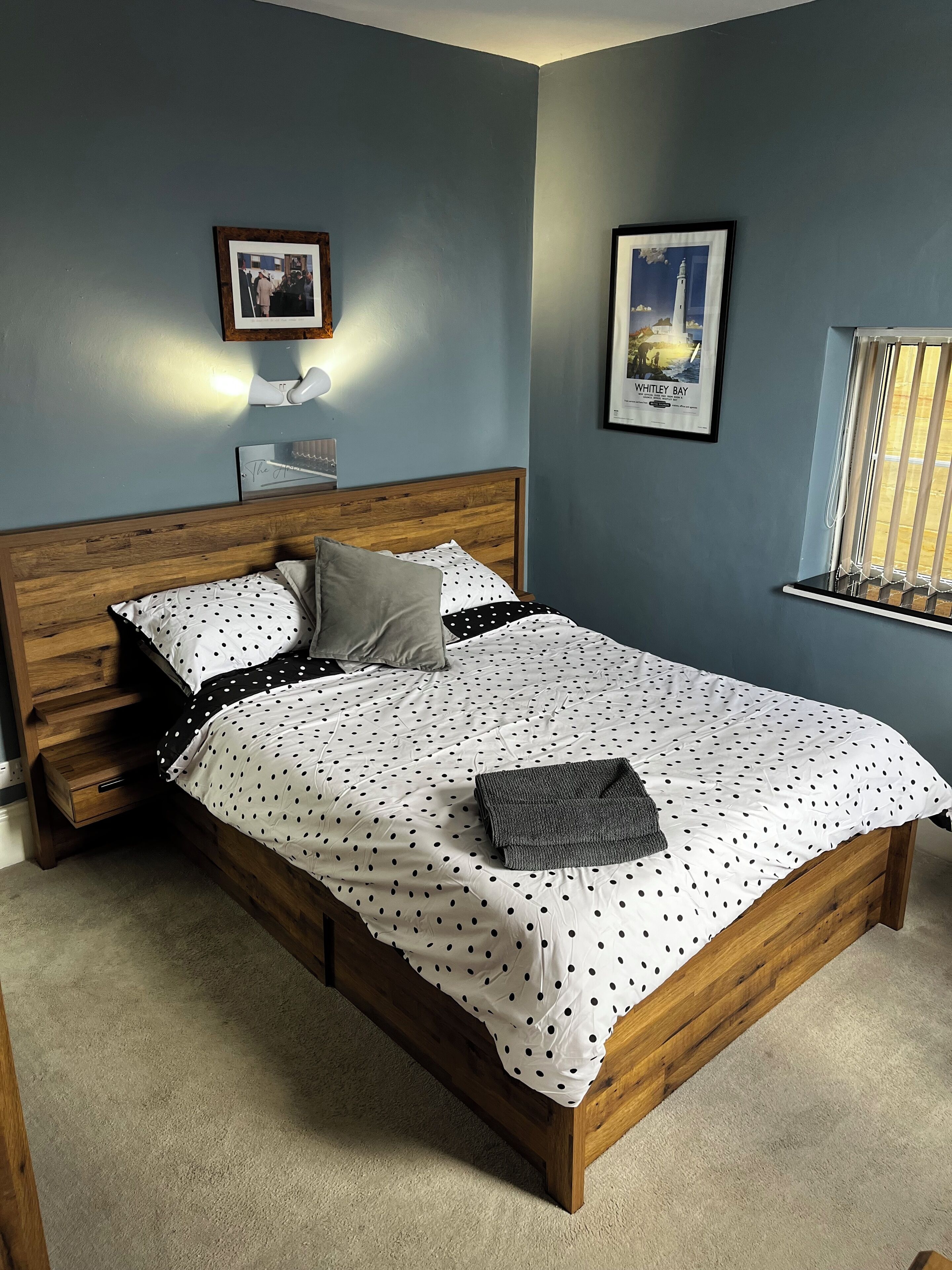 A bedroom with a wooden bed, a nightstand, a framed picture on the wall, and a window with blinds.