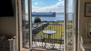 Et værelse med en stor skydedør, der fører til en balkon med et bord og stole, udsigt til et skib, der hedder HOËGH AUTOLINERS, og et TV ophængt på væggen.