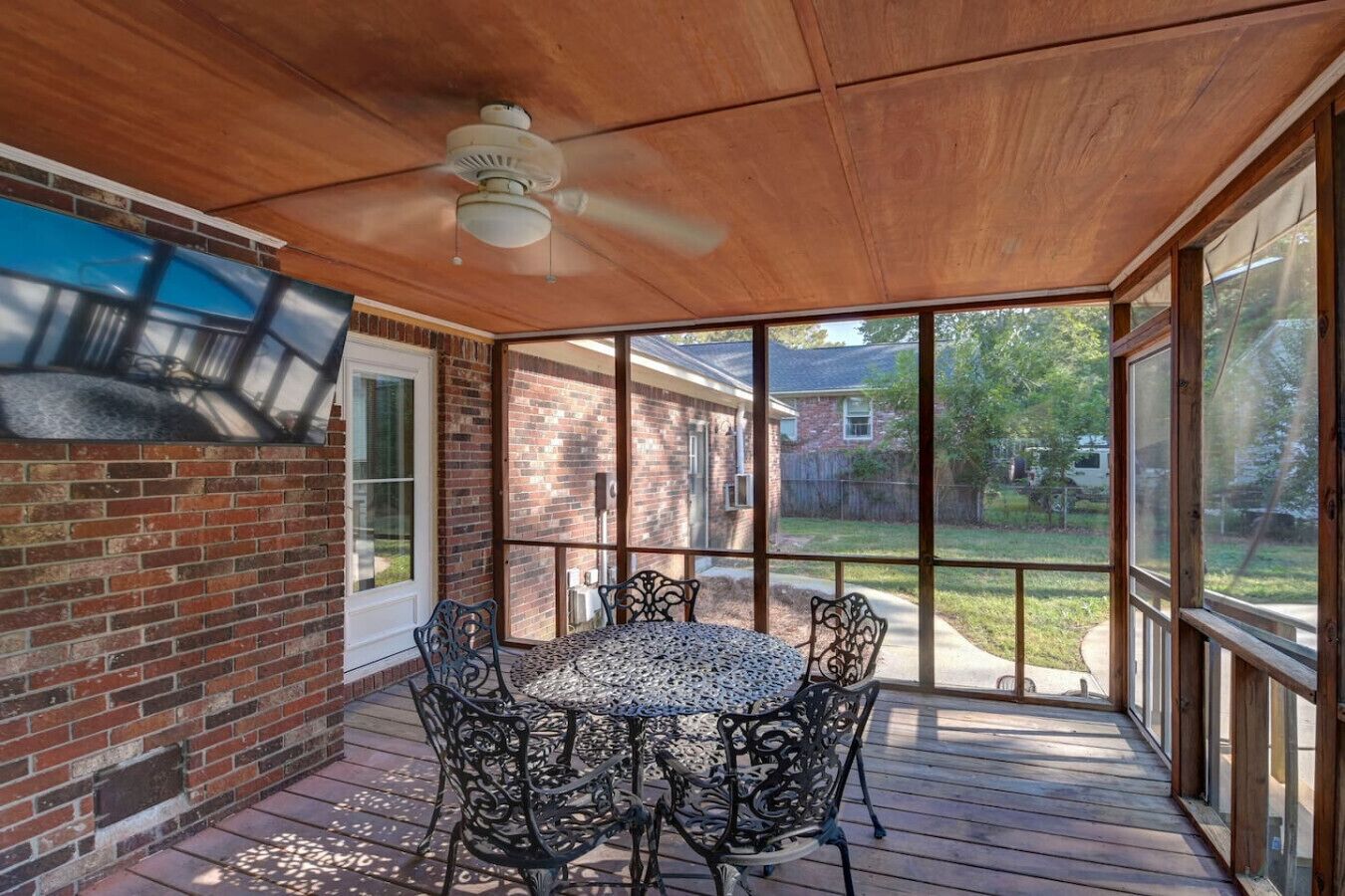 Een overdekt terras met een ronde tafel en stoelen, een plafondventilator en grote ramen met uitzicht op de achtertuin.