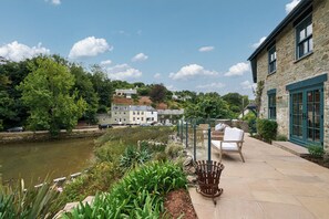 A riverside patio with outdoor furniture, a stone building with green shutters, and a backdrop of houses and trees. - Luxury 3-Bed House with Boathouse on the Estuary–Outdoor Kitchens & Hot Tubs for (Salcombe)
