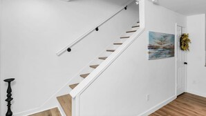 A staircase with white walls and wooden steps, a painting on the wall, and a door with a wreath. - Stay West Side | 2 BR | Minutes from Downtown (West Columbia)