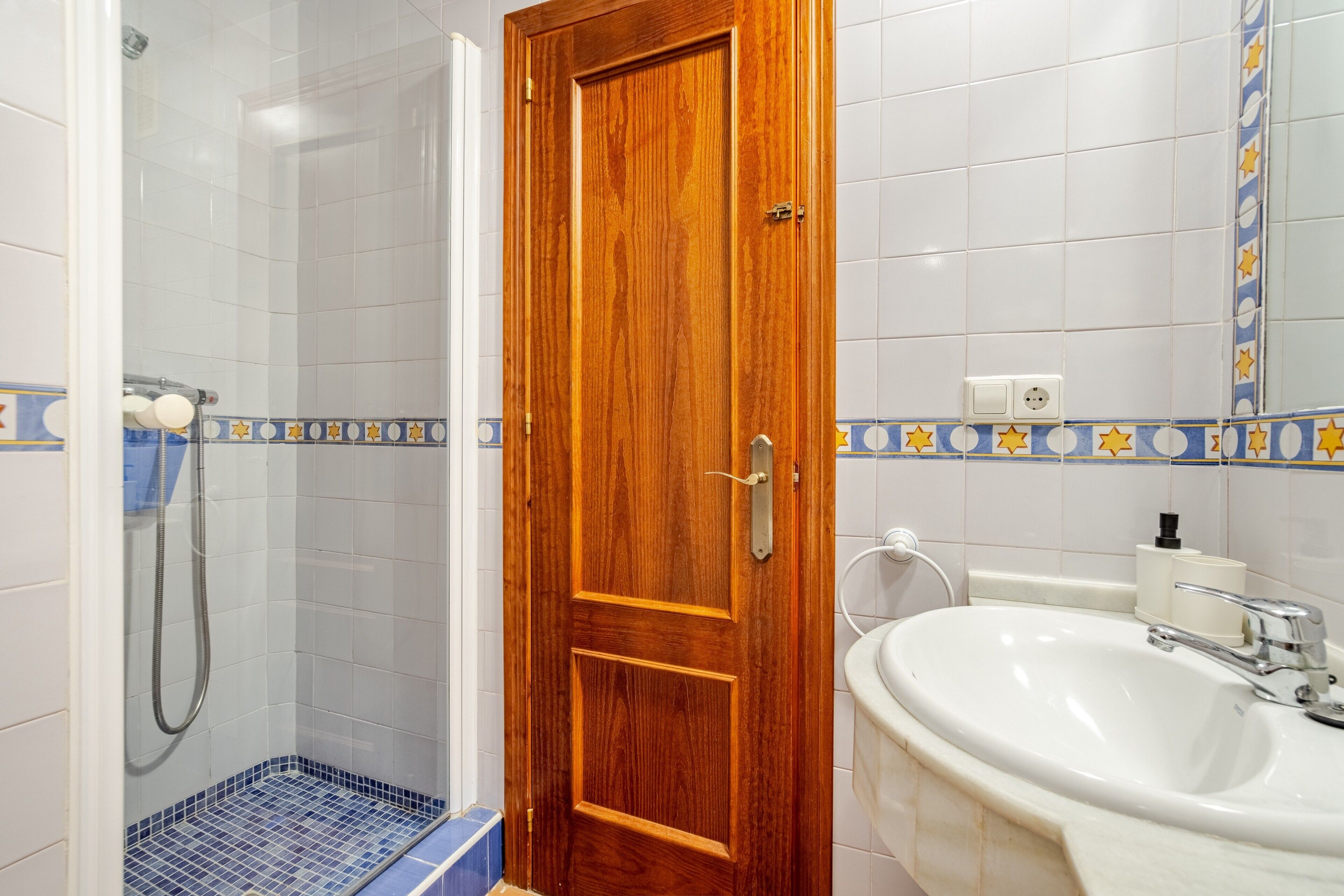 A bathroom with a wooden door, a shower, a sink, and a tiled floor.