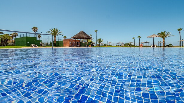 Una piscina con agua azul cristalina, rodeada de palmeras y un pabellón.