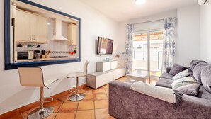 Una sala moderna con sofá, comedor con mesa y sillas, cocina con armarios y televisor, y balcón con vistas.